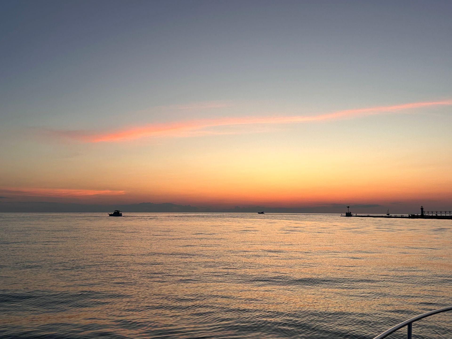 Sunset over a calm ocean. Orange and pink hues in the sky, a boat and a pier in the distance.