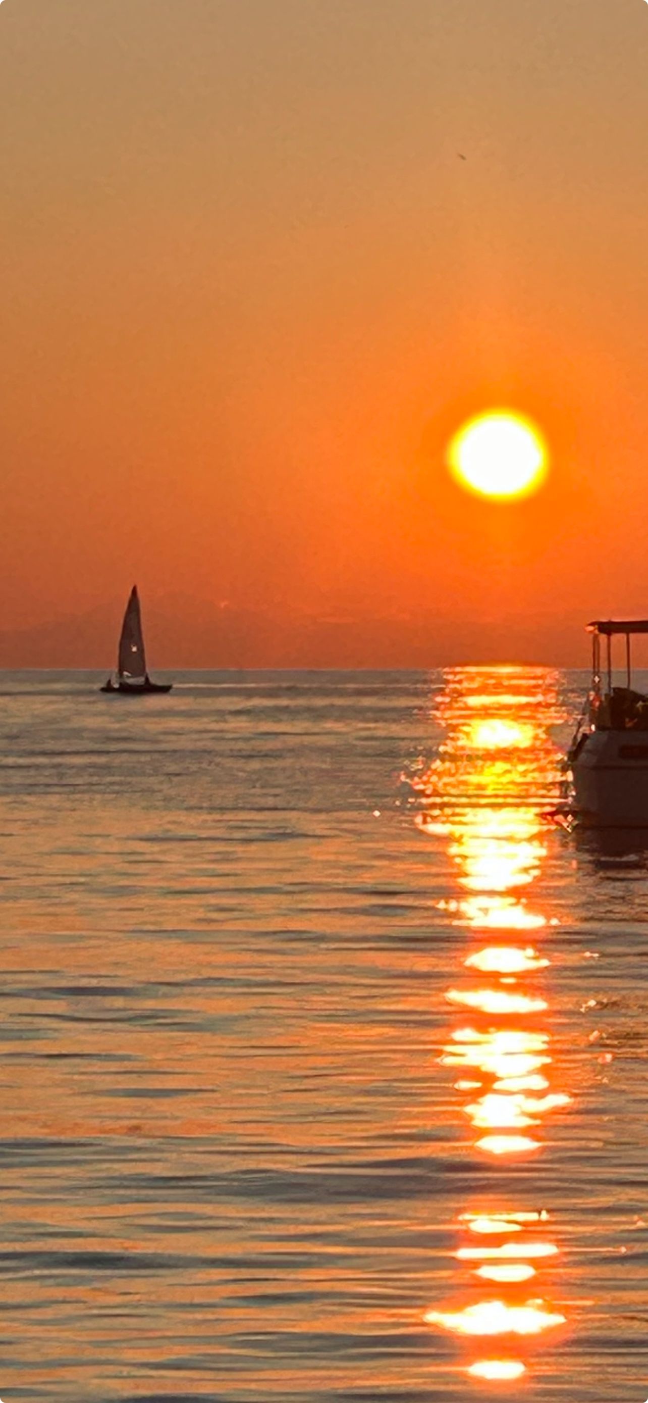 Sunset over ocean, with the sun's reflection on water, a sailboat and a boat. Orange hues.