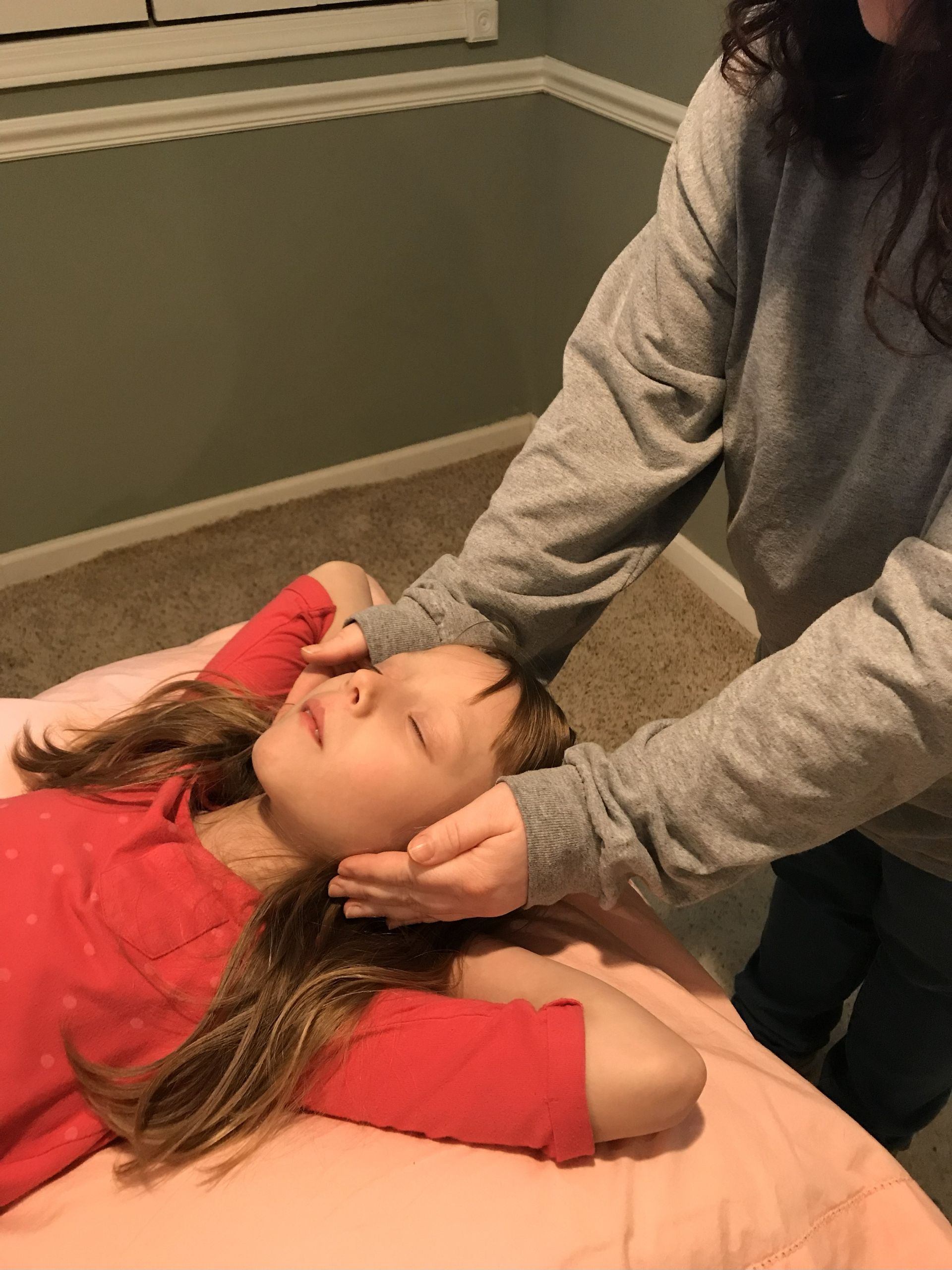 Person gently holds a child's head, possibly administering therapy. Child lies with closed eyes on a bed.