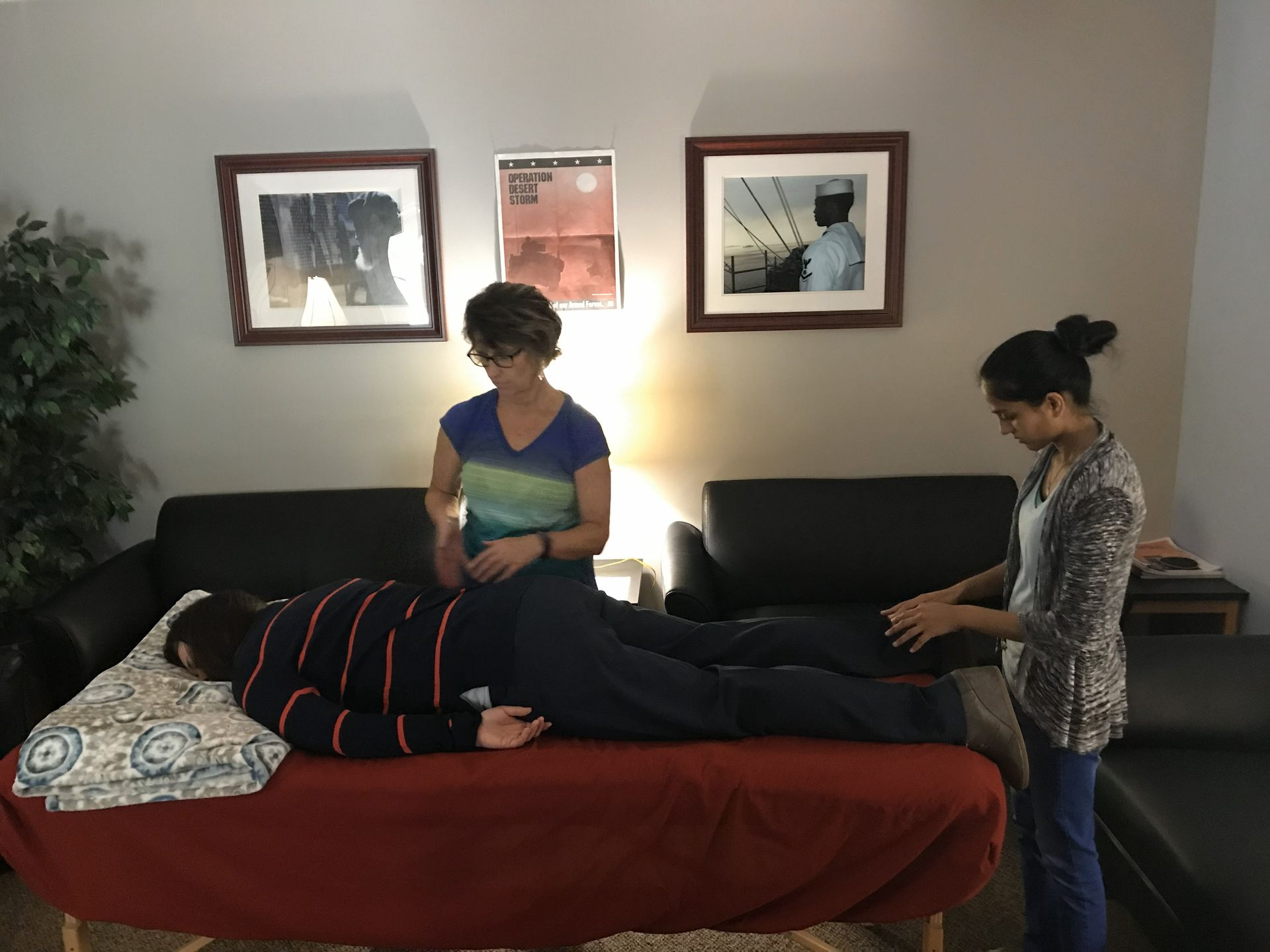 Two people giving a massage to a person lying on a table. The room has framed art and a couch.