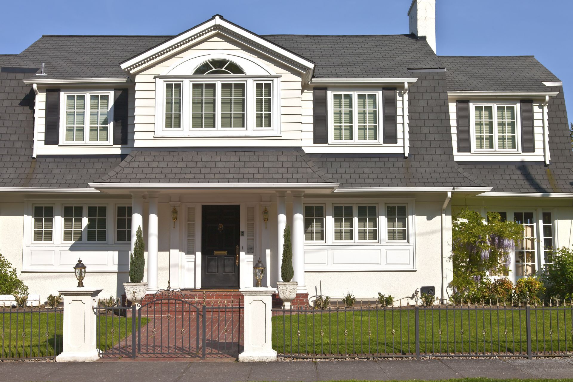 White two-story house with dark gray roof, black shutters, and brick porch. Green lawn and black fence in front.