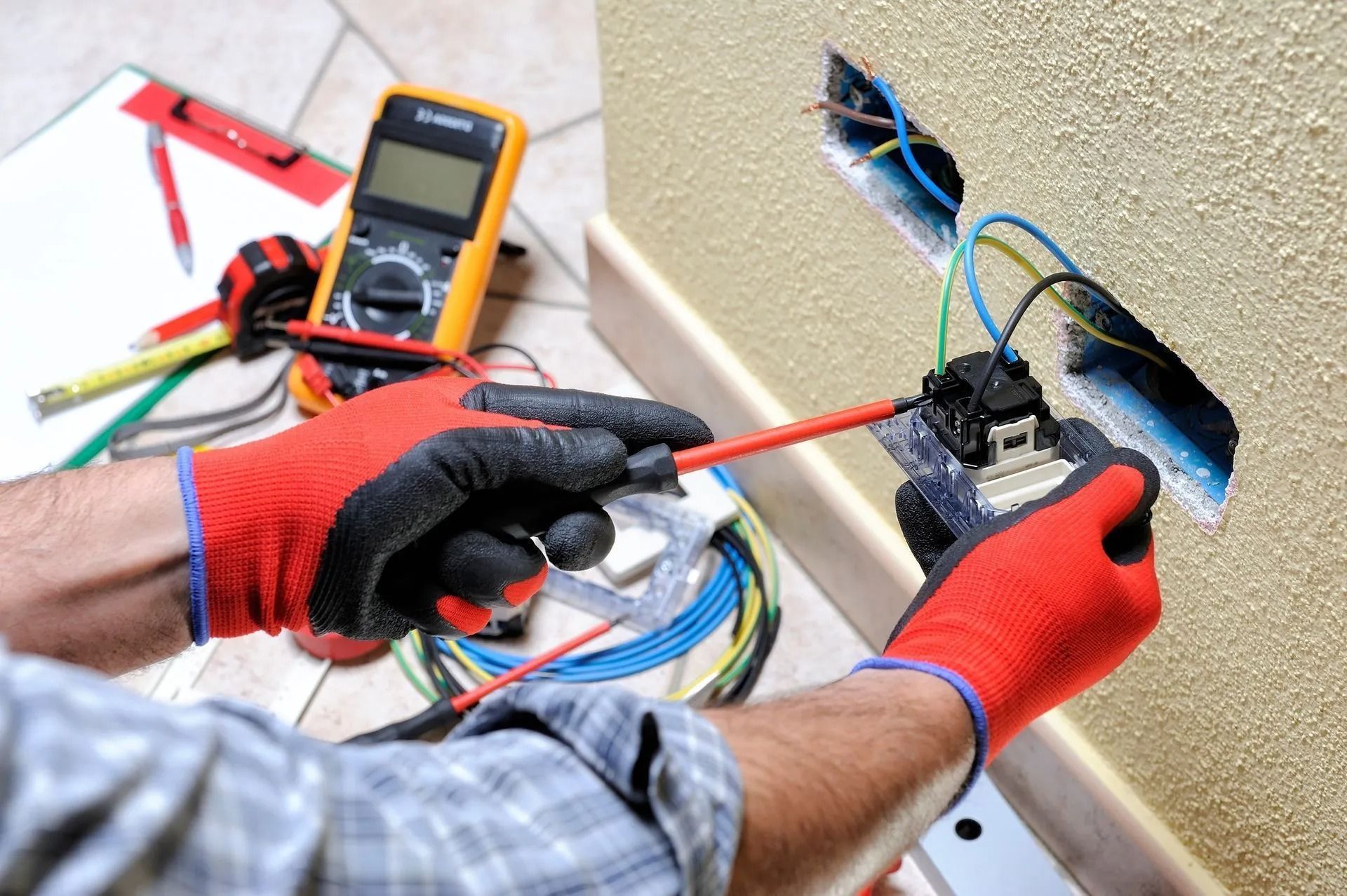 Electrician installing an electrical outlet, wearing red gloves, tools in background.