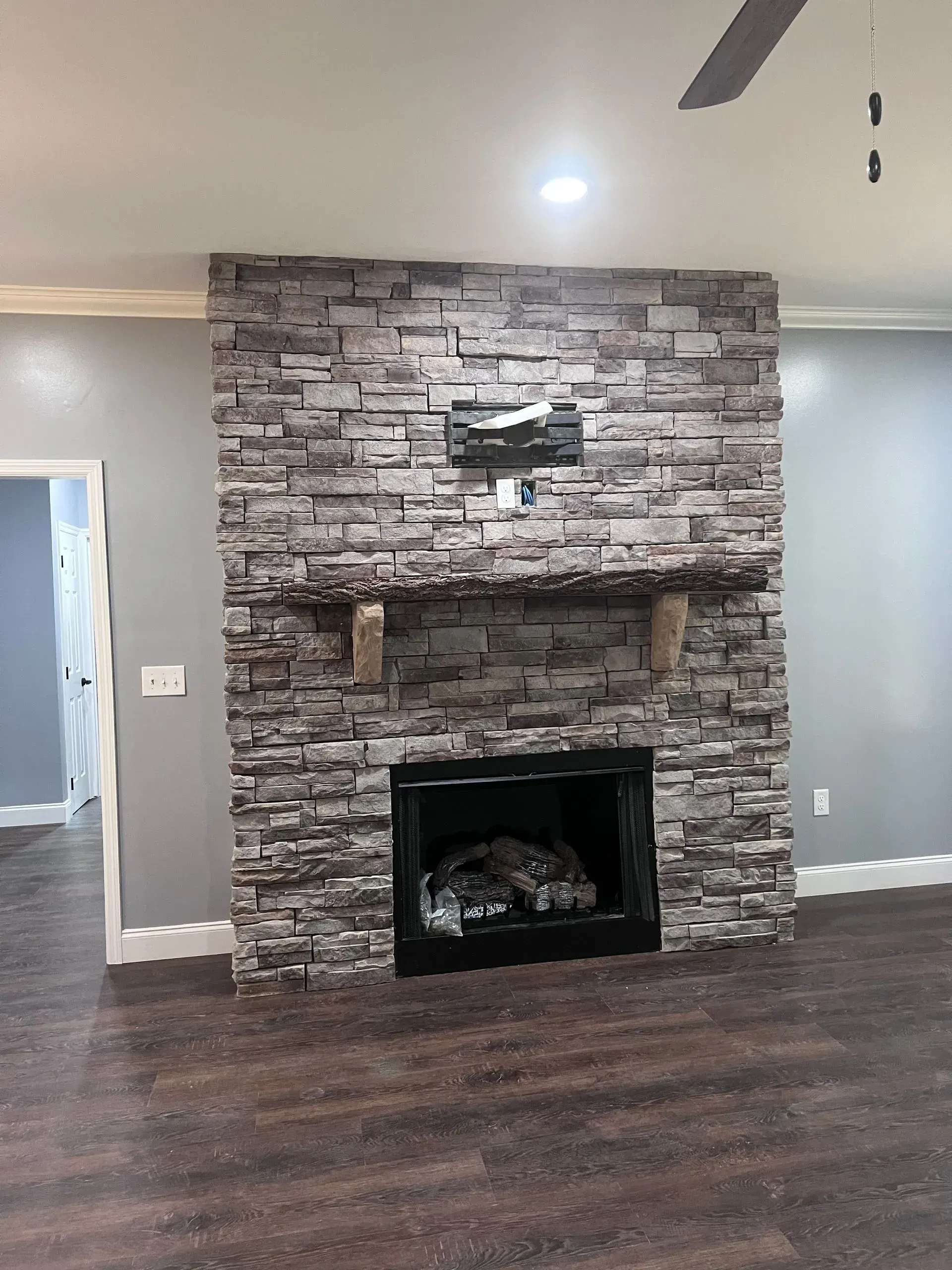 Fireplace with gray stone facing, black hearth, and wooden mantel in a room with dark wood flooring and gray walls.