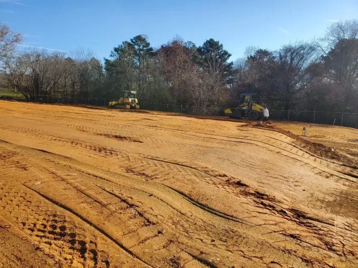 Two bulldozers leveling a dirt field near trees on a sunny day.