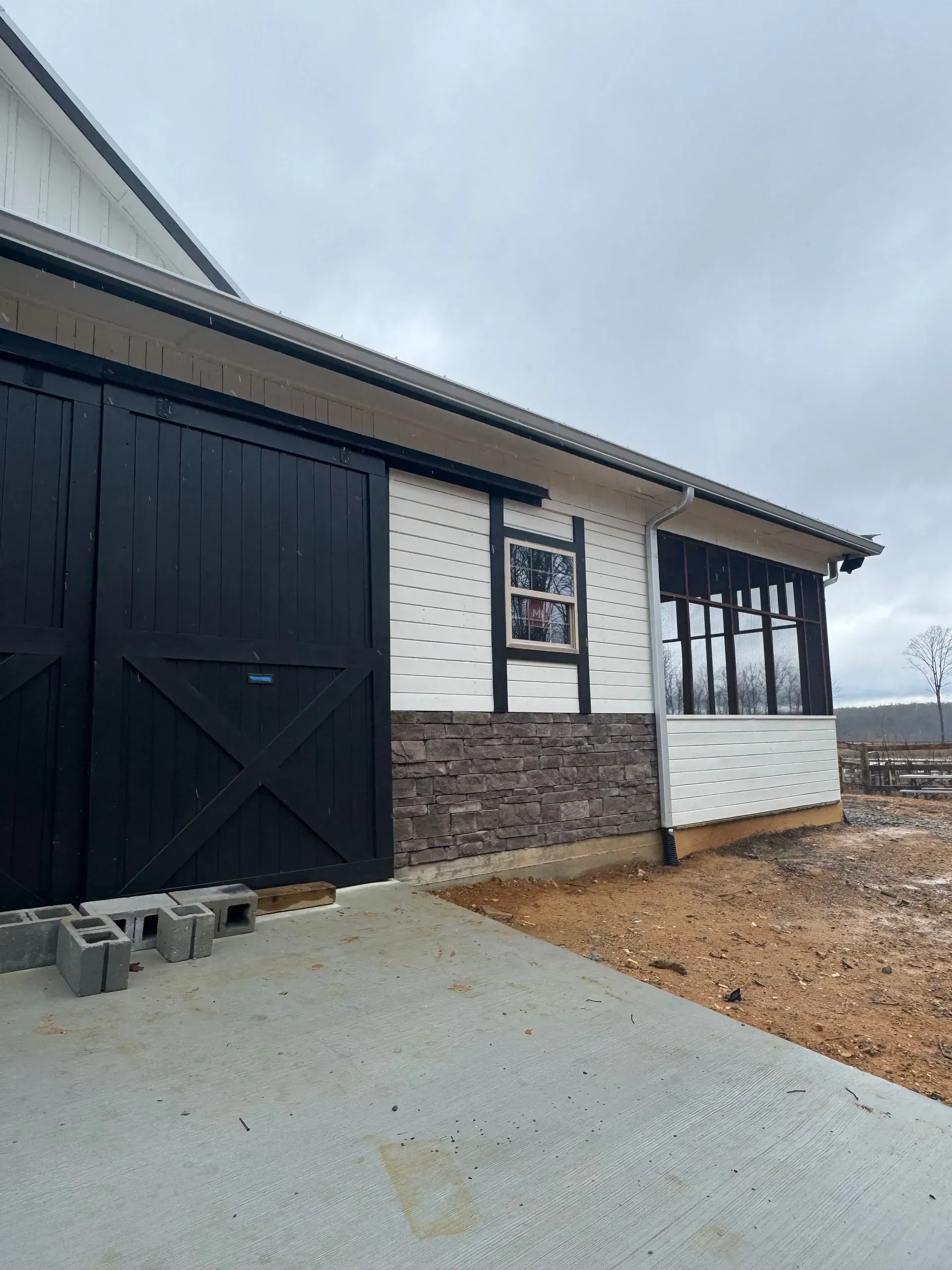 Black barn with sliding doors, white siding, and brown brick. Gray sky.