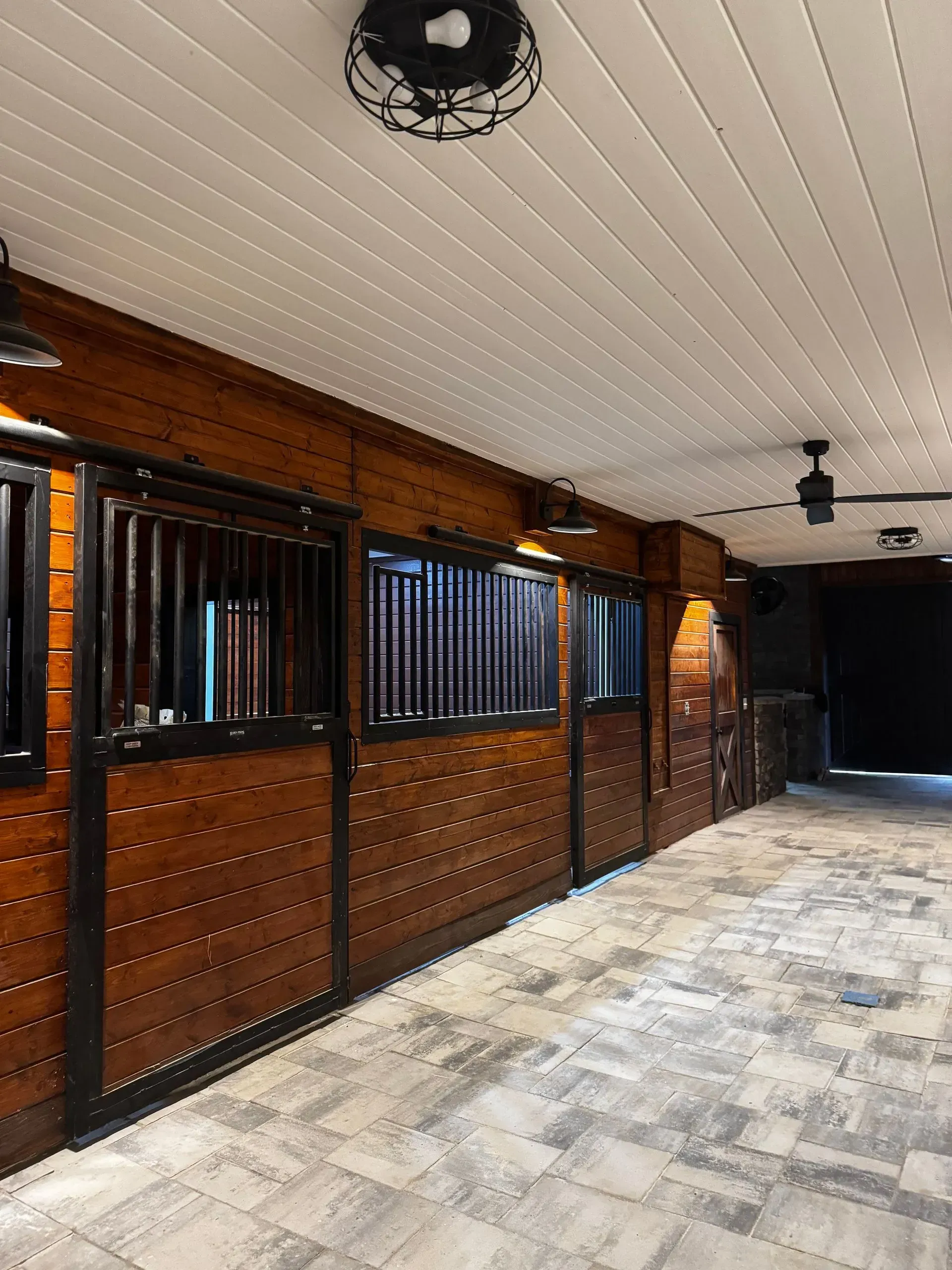 A row of wooden horse stalls with black metal grilles in a barn with a tiled floor and ceiling lights.