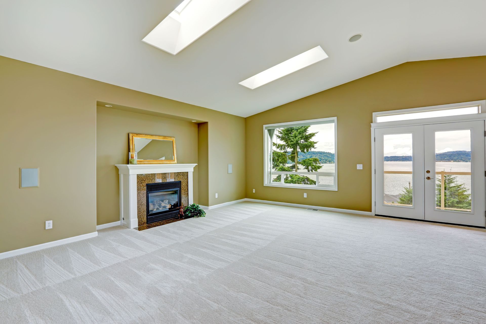 Empty living room with fireplace, window, and doors overlooking a lake, beige walls and carpet.