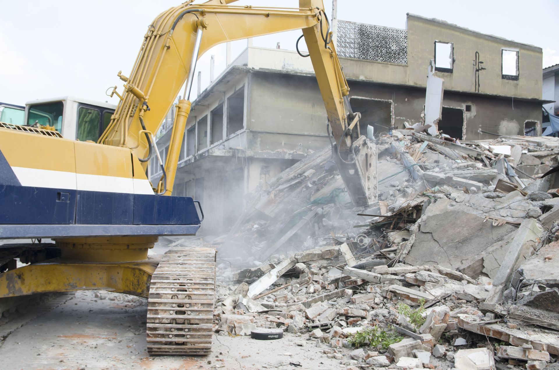 Yellow excavator demolishing a building, creating a pile of debris and dust.