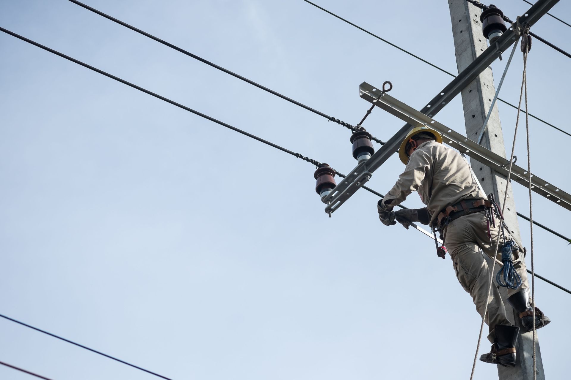 Lineman working on power lines attached to a utility pole against a blue sky.