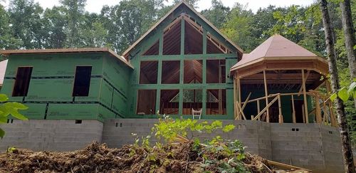 House under construction in a wooded area, with timber frame and green sheathing visible.