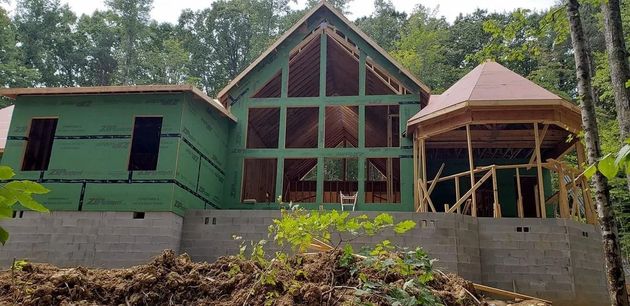 House under construction, with a large paneled window area, gazebo, and green sheathing. Surrounded by trees.