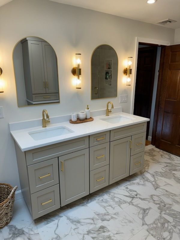 A double vanity with grey cabinets, gold hardware, white marble countertop, arched mirrors, and gold wall sconces.