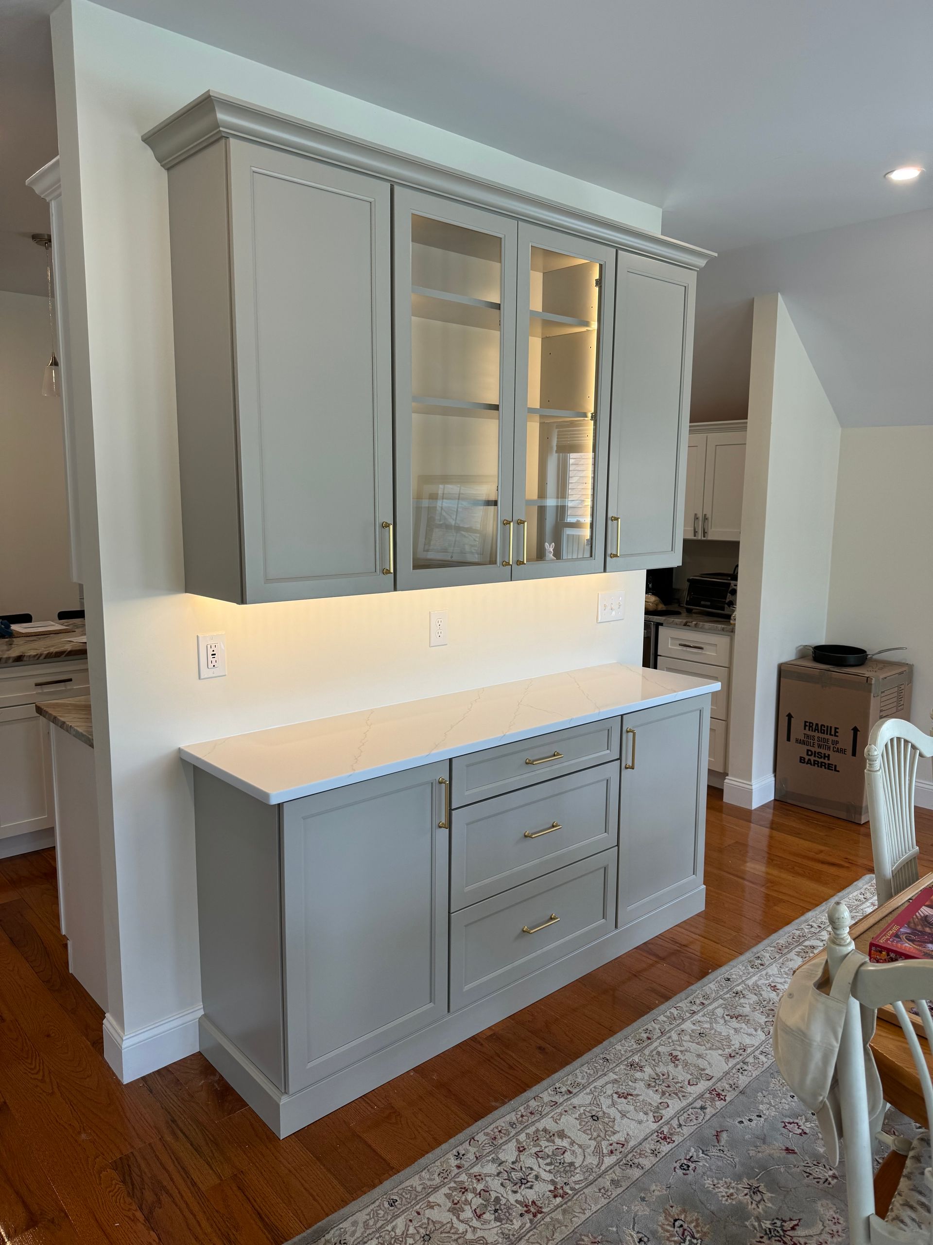 White built-in kitchen cabinet with glass-front upper doors, lit backsplash, and lower drawers in a bright room