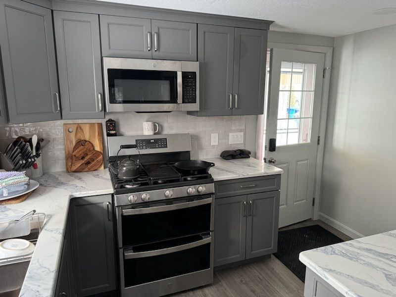 Modern kitchen with gray cabinets, stainless steel appliances, marble-patterned countertops, and a glass-paneled door.