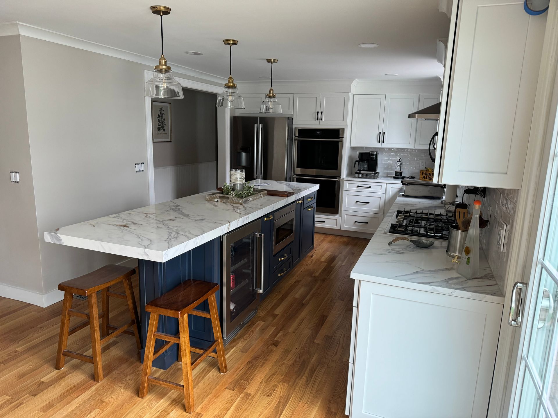 Modern kitchen with marble island, dark stools, stainless fridge, and white cabinets on wood floors.