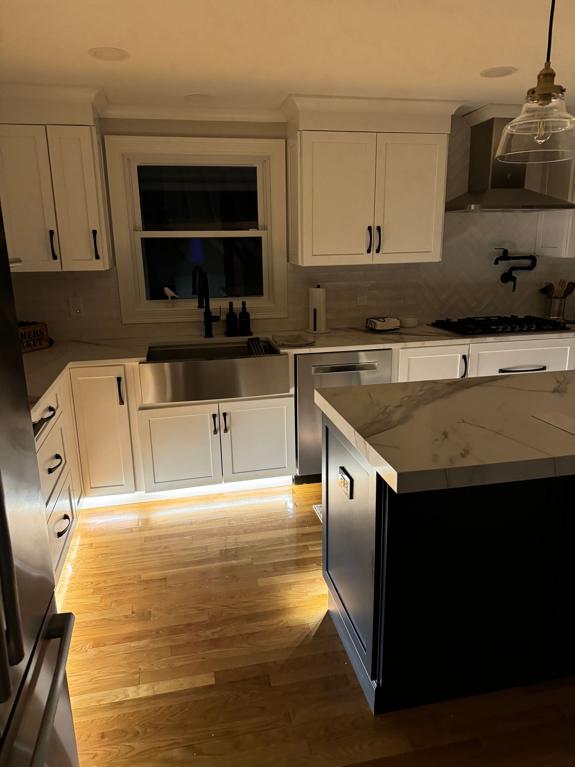 Dim kitchen with white cabinets, black island, pendant light, and under-cabinet lighting on hardwood floor.