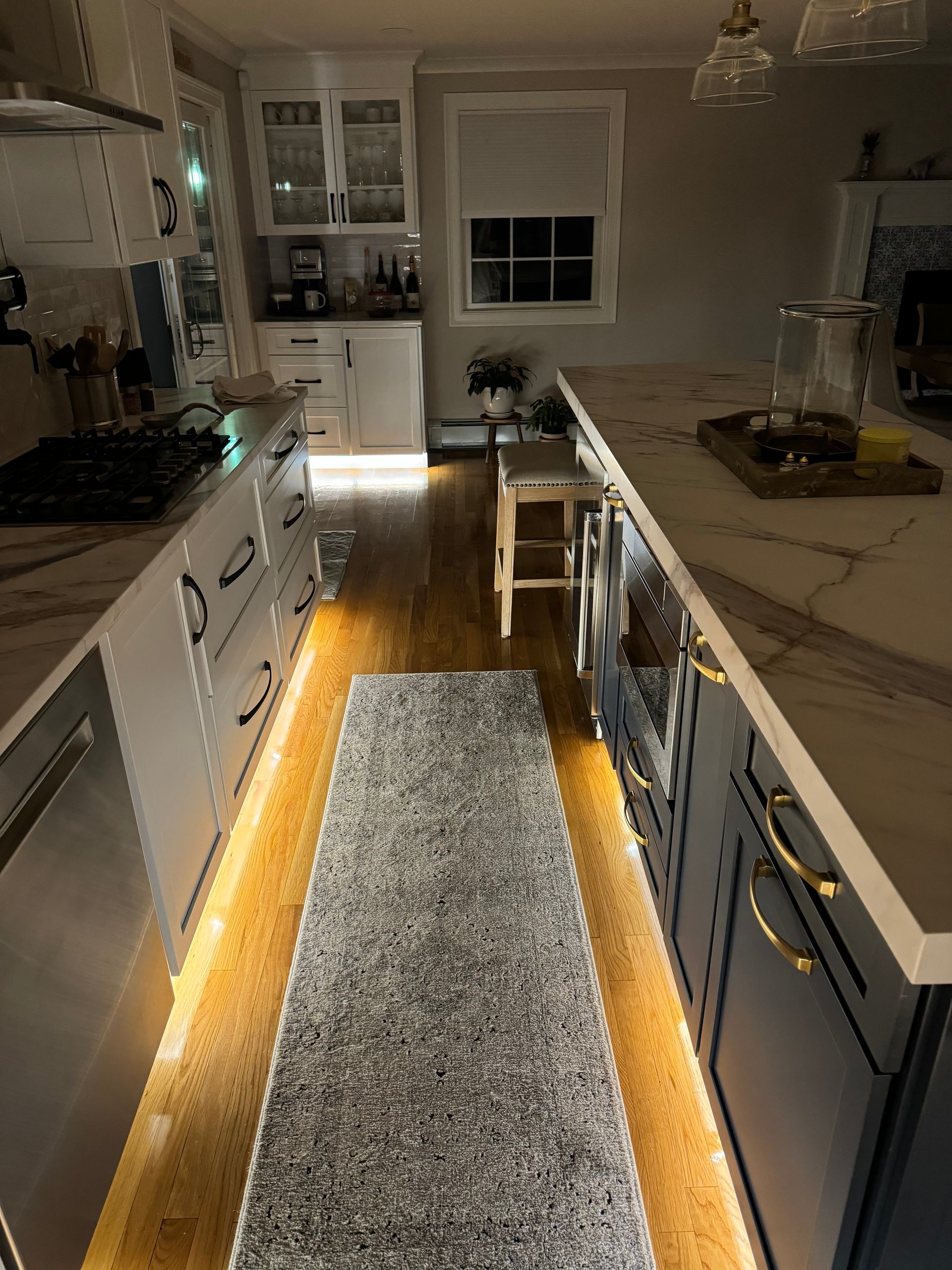 Narrow galley kitchen with white cabinets, a central rug, and warm under-cabinet lighting on both sides.