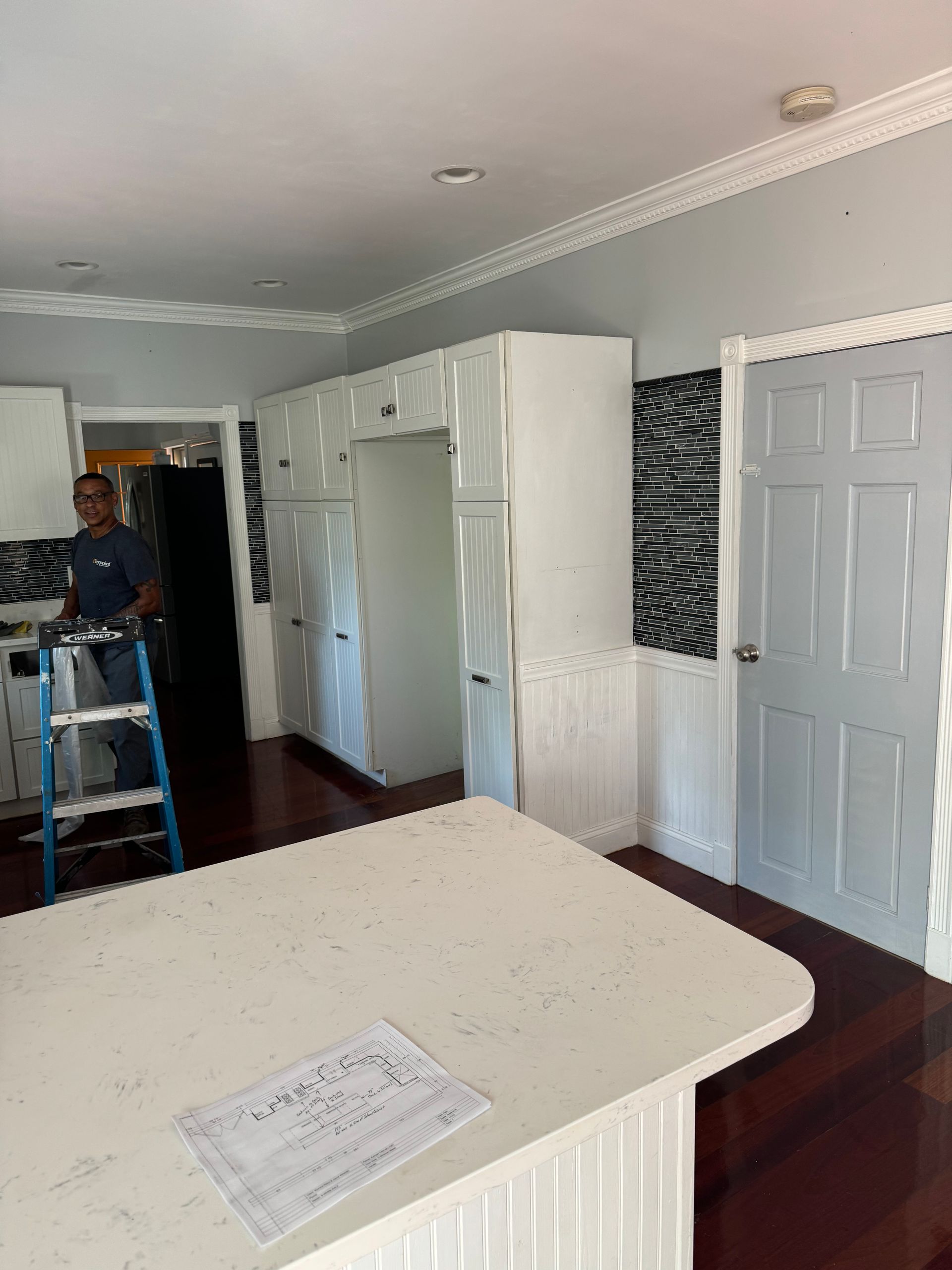 Bright kitchen with white cabinets, gray walls, and a person standing on a blue step ladder near the doorway