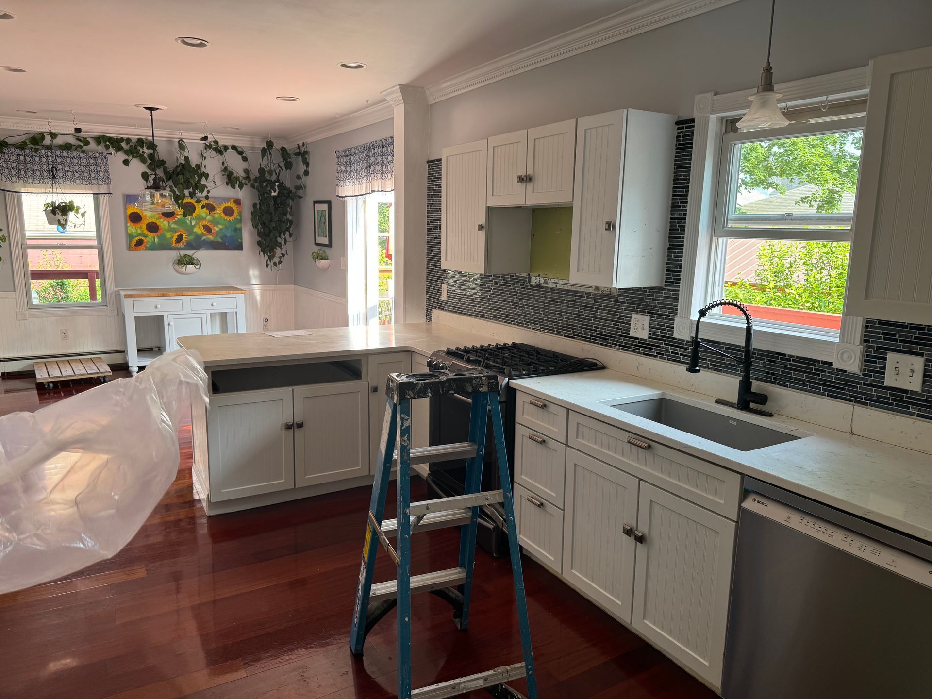 Kitchen under renovation with white cabinets, dark counters, ladder, and a covered island near a sunny window.