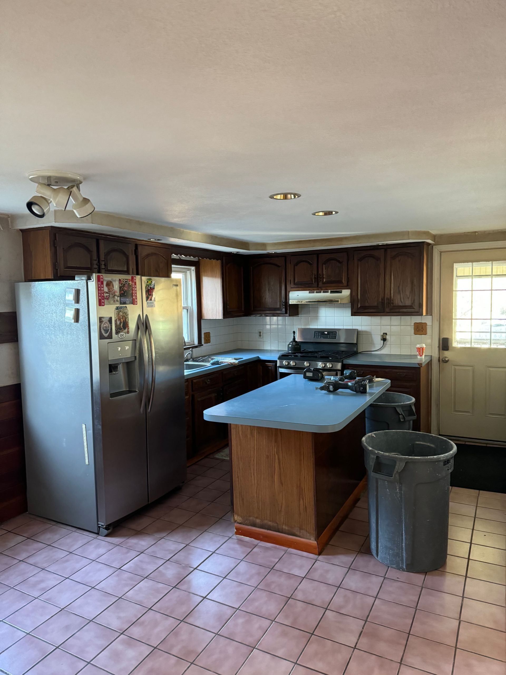 Small kitchen with stainless fridge, dark cabinets, central island, tiled floor, and trash can by the window.