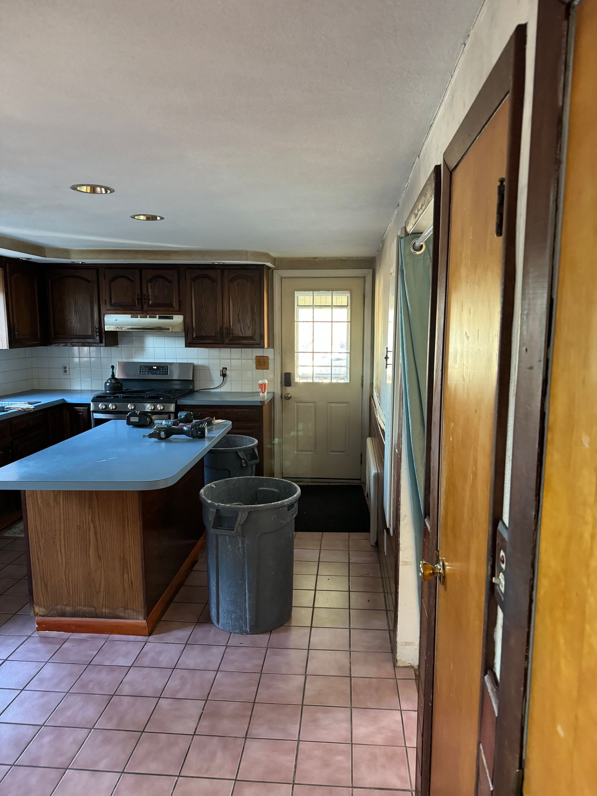 Narrow kitchen with tile floor, dark wood cabinets, island, trash can, and a door at the far end