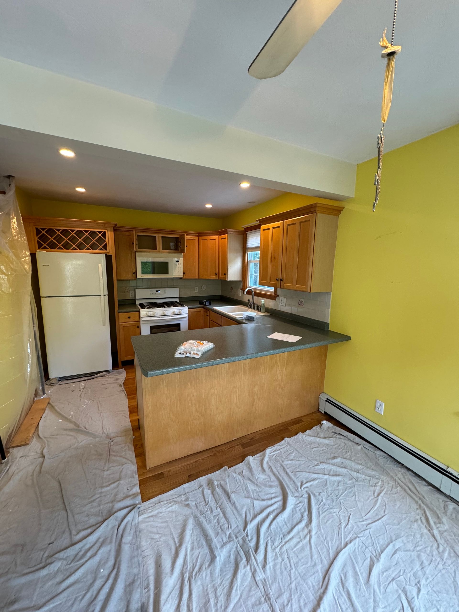 Small kitchen with green walls, wood cabinets, white appliances, and a taped plastic sheet on the floor.