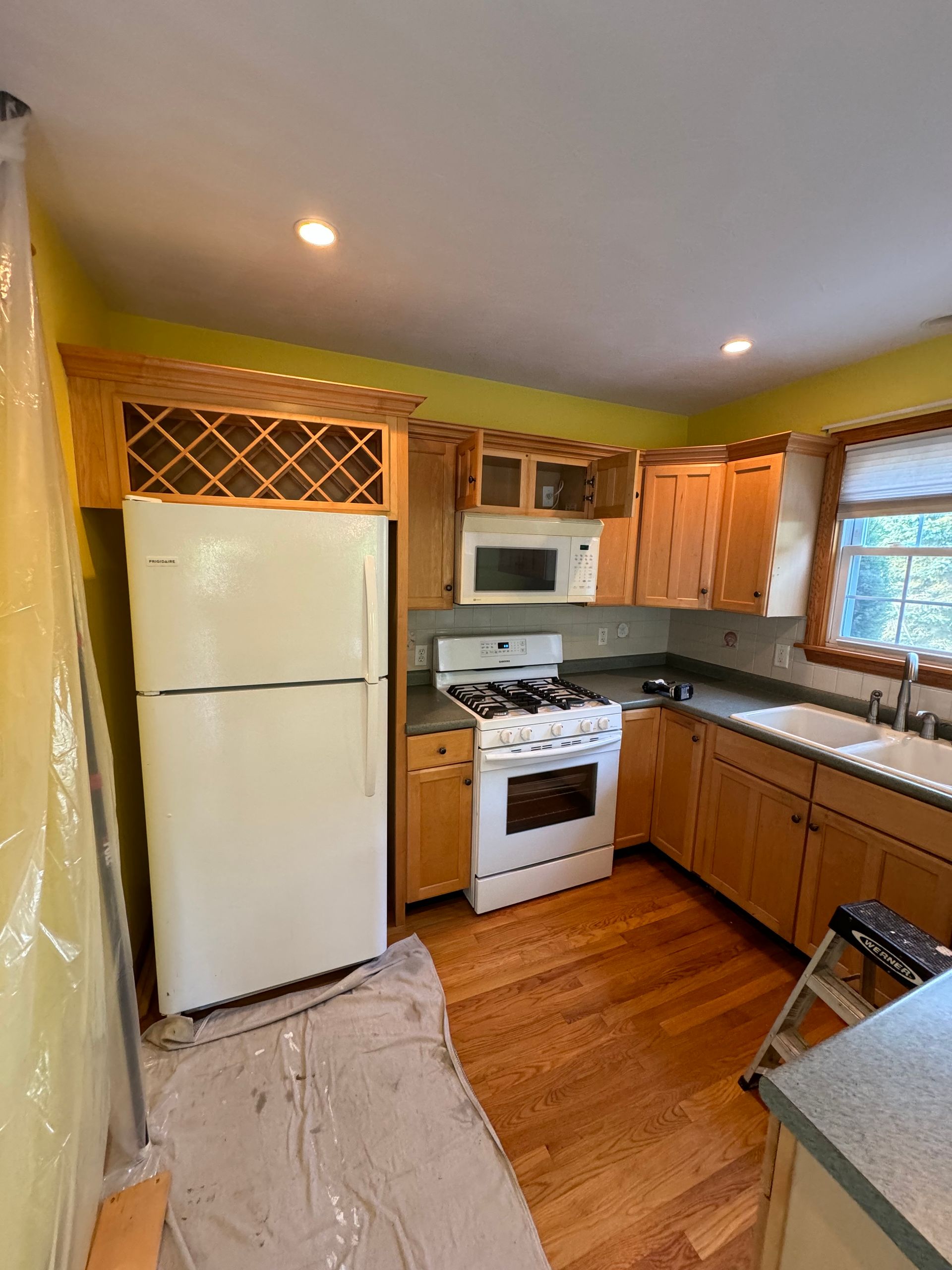 Small kitchen with white appliances, wood cabinets, yellow walls, and a rug-covered floor under construction.