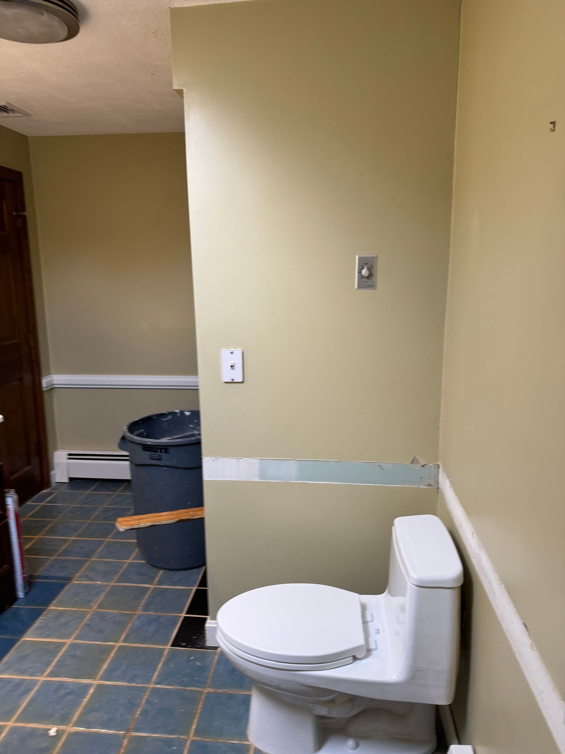 Small bathroom with a toilet in the foreground, beige walls, blue tiled floor, and a sink area in back.