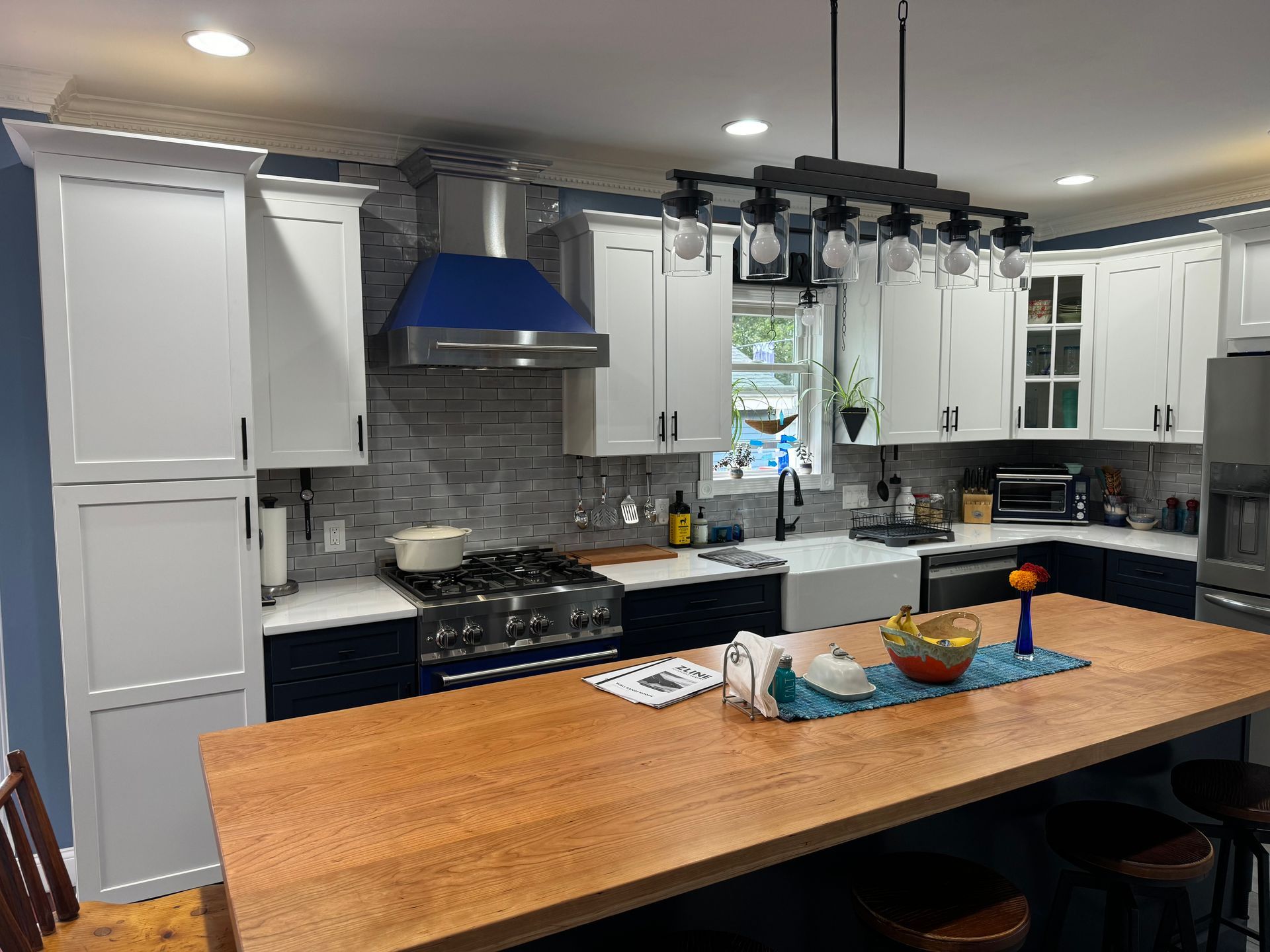 Modern kitchen with white cabinets, blue range hood, butcher block island, dark blue base cabinets, and gray backsplash.