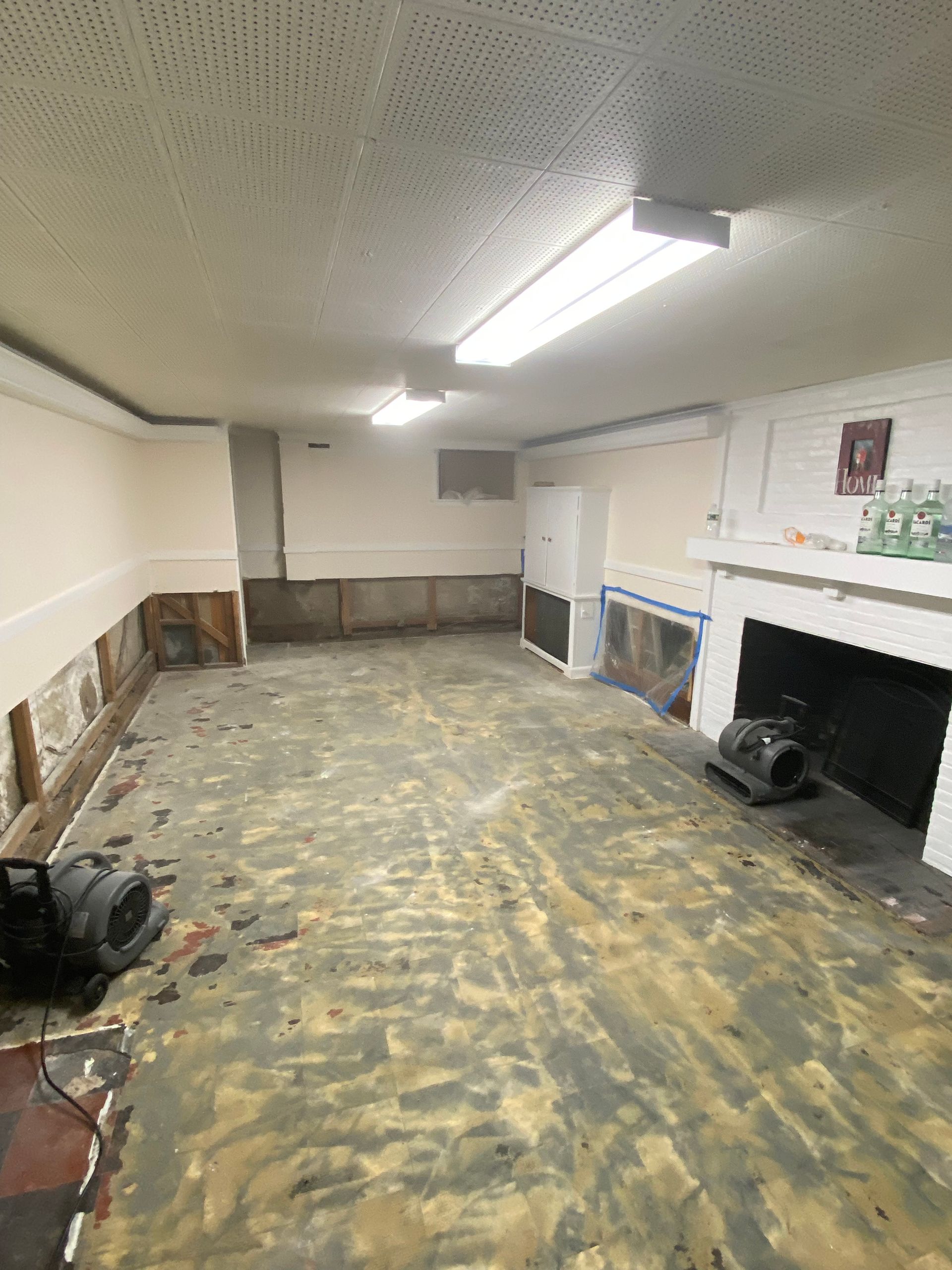 Empty basement room with speckled floor, white walls, fluorescent lights, and a fireplace on the right.