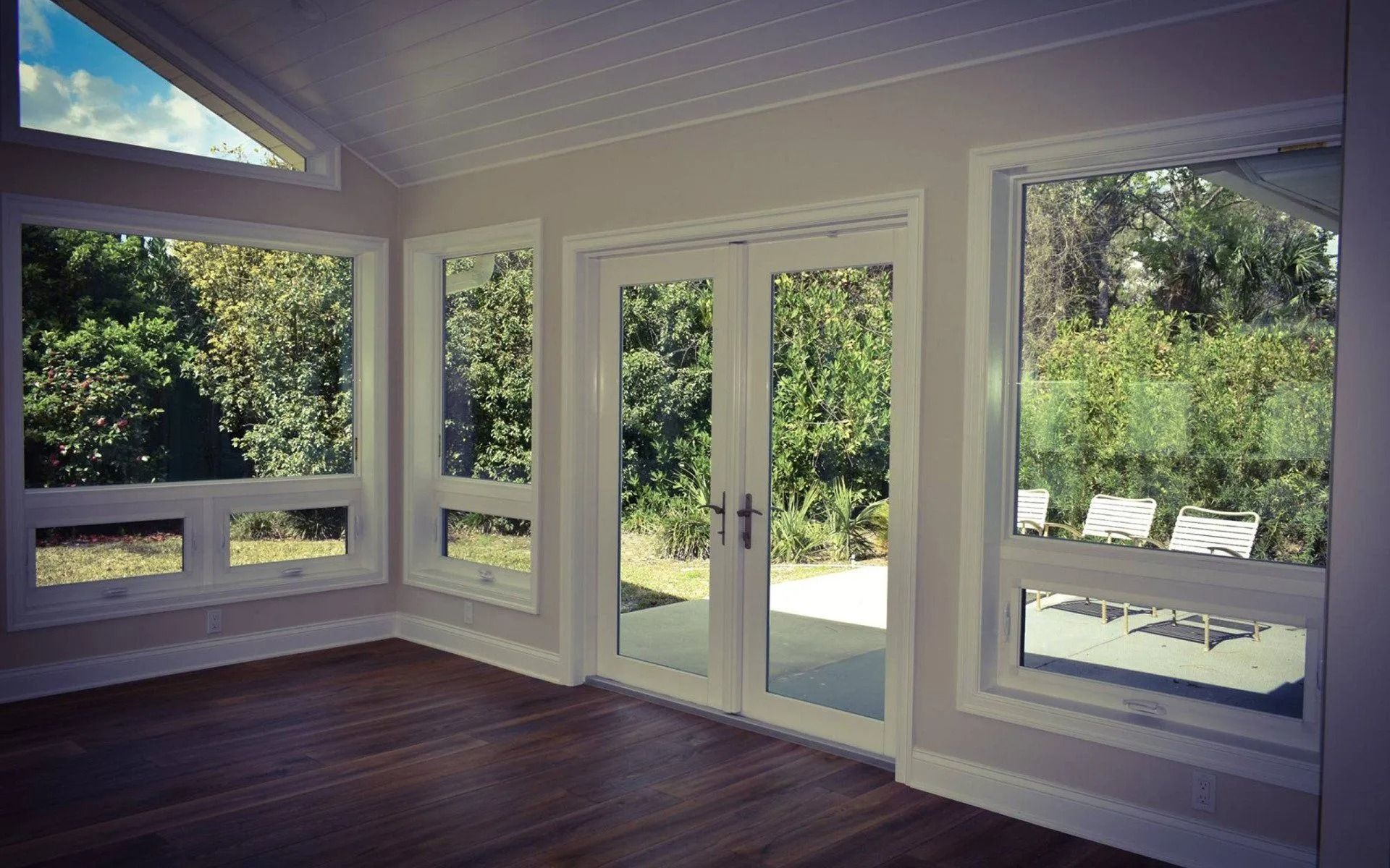 Interior room with large windows and glass doors overlooking a green yard, hardwood floors, white trim.