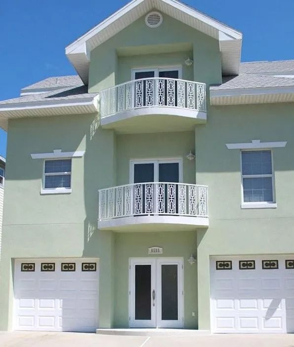 Two-story green house with white balconies, garage doors, and windows; clear blue sky.