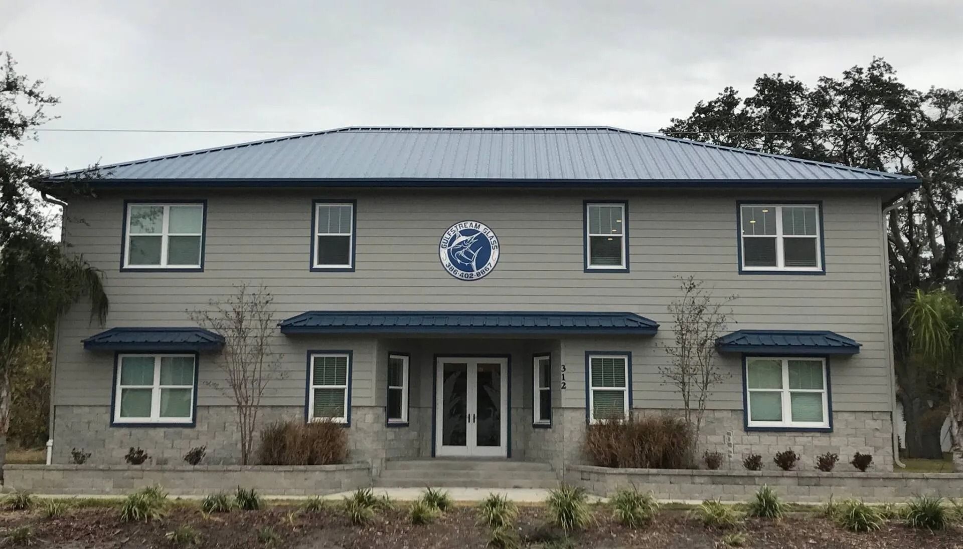 Two-story building with gray siding, blue metal roof, and a logo in the center.