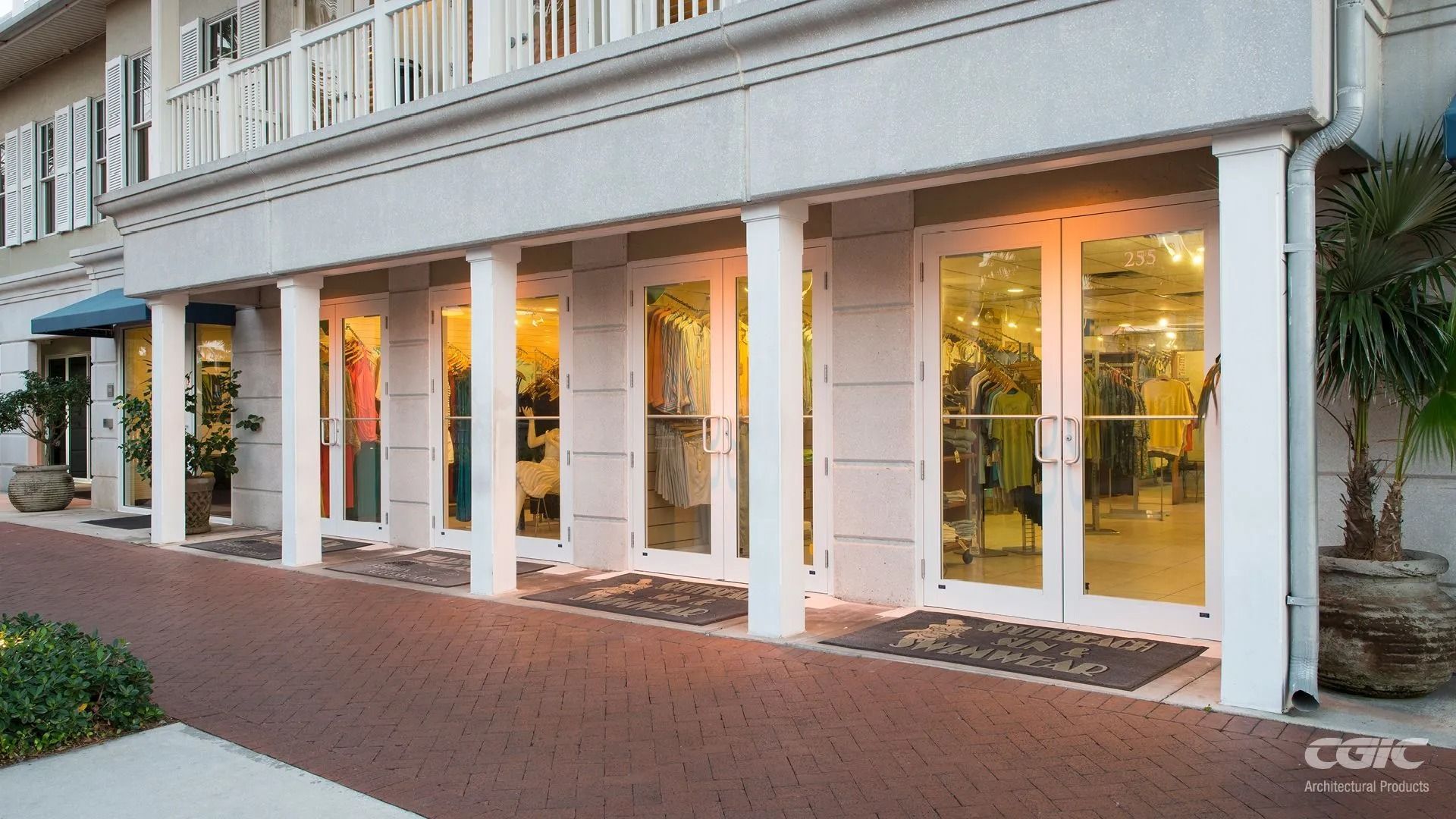 Storefront with white columns, glass doors, and clothing displayed inside. Exterior with brick walkway and a potted palm.