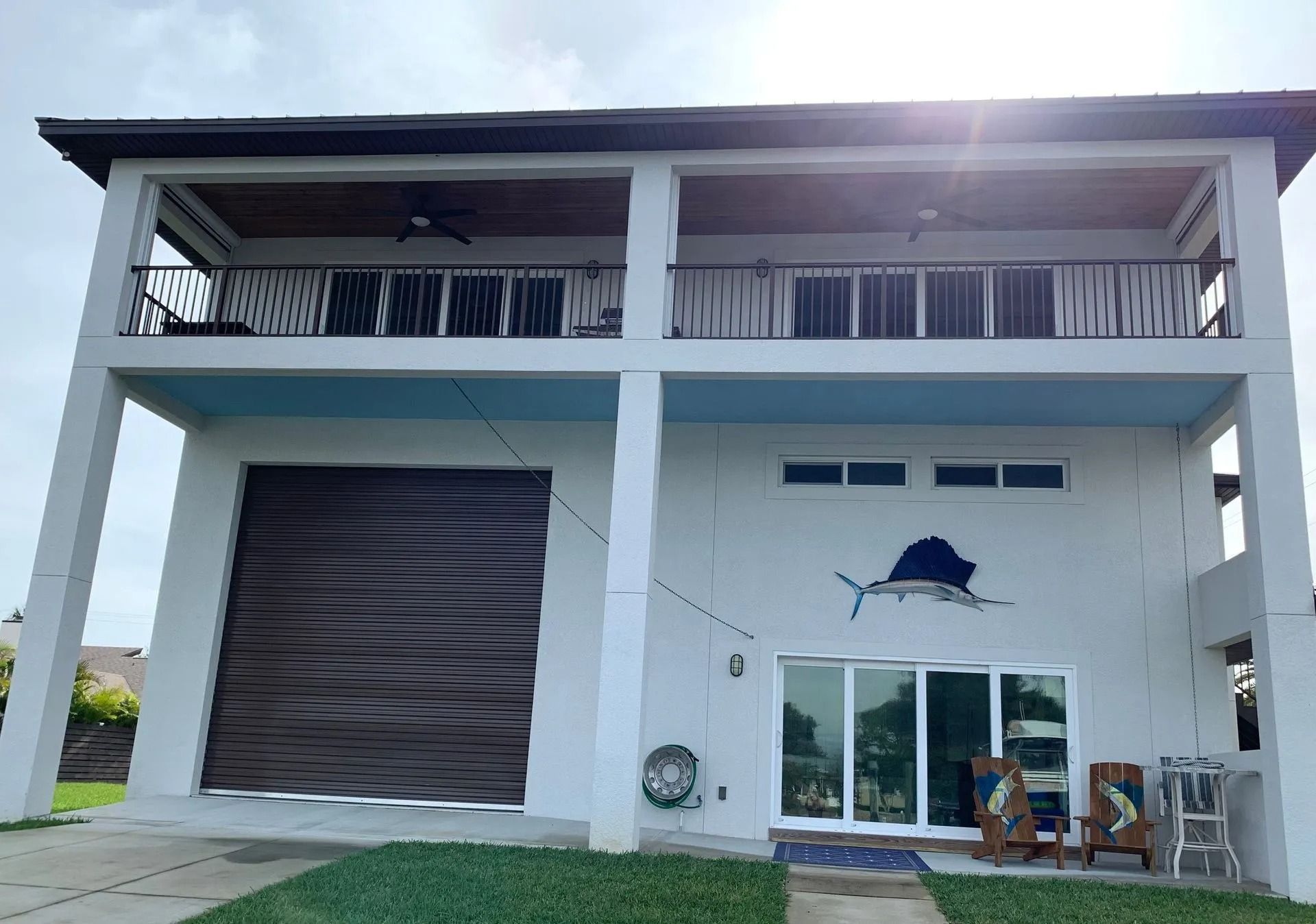 A two-story white house on stilts with a dark rolling garage door, a second-floor balcony, and a sailfish wall decoration.
