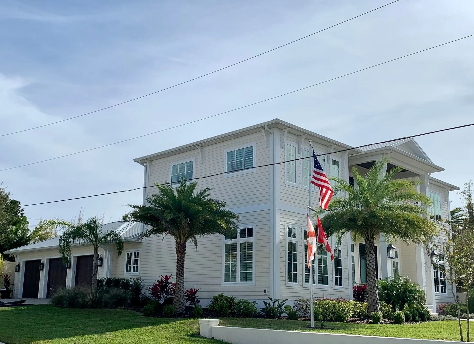 A large, two-story, light-colored house with a three-car garage, palm trees, and an American flag in the front yard.