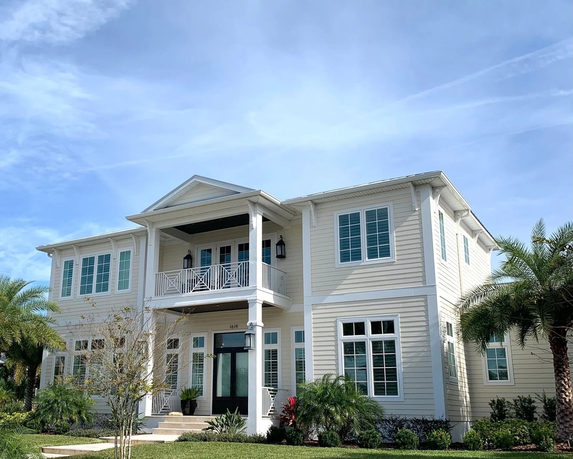A two-story, cream-colored house with a front balcony, dark front door, and symmetrical windows under a bright blue sky.