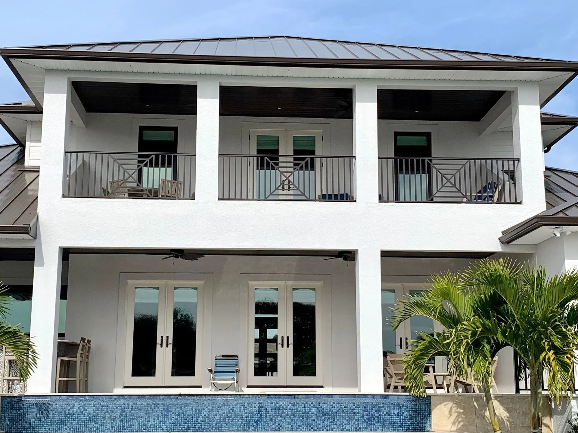 A two-story white house with a metal roof, balconies, large glass doors, and a blue tiled pool in the foreground.