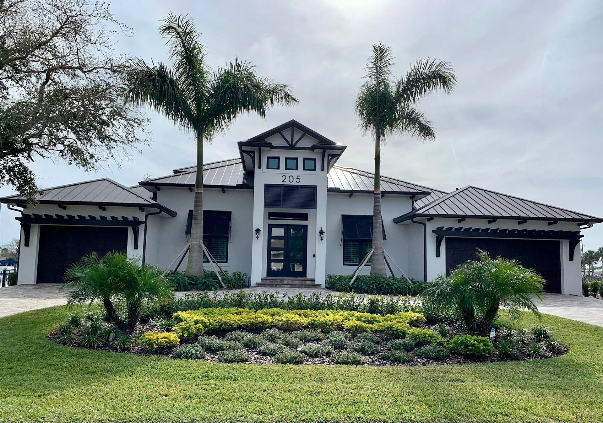 A modern, white single-story home with a dark roof and two garages, featuring a landscaped front garden and palm trees.