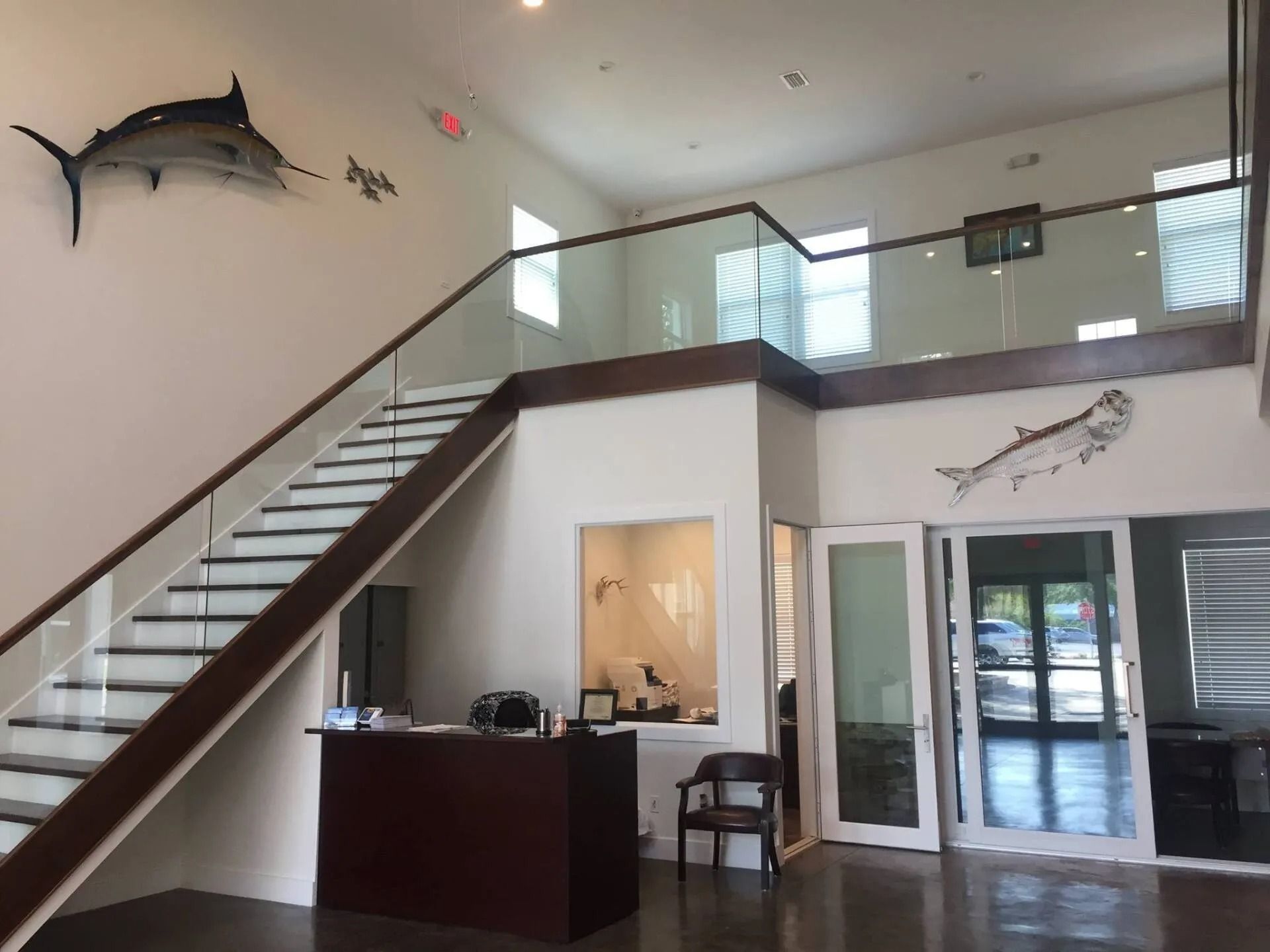 A bright office lobby with a modern staircase, a reception desk, glass railings, and wall-mounted fish decor.