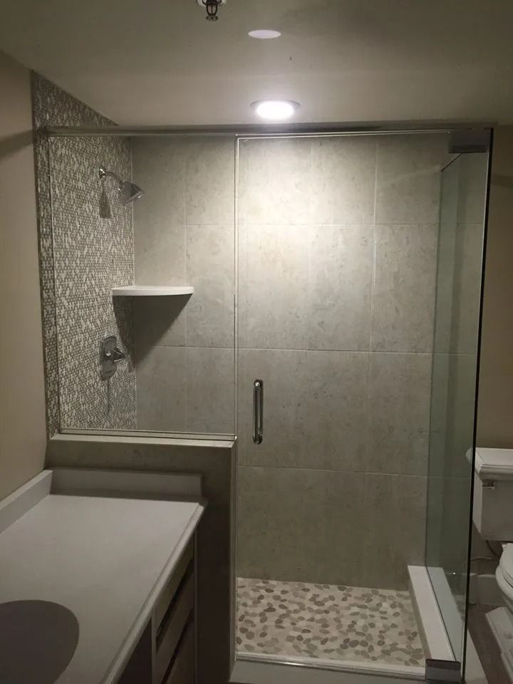 A modern walk-in shower with a glass door, pebble-stone floor, patterned tile wall, and a built-in corner shelf.