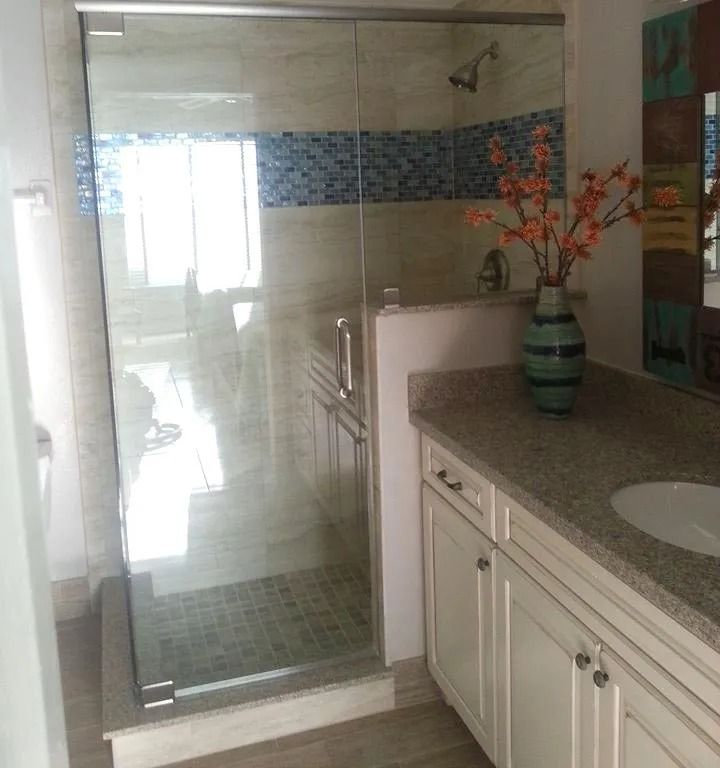 Bathroom with glass shower, white vanity, and granite countertop. Blue and neutral-toned tile walls. Orange flowers in a vase.