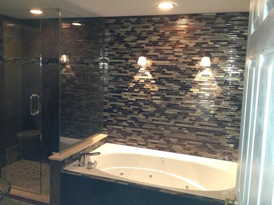 Bathroom with tub and shower; mosaic tile wall, two sconces, dark cabinetry, and glass shower door.