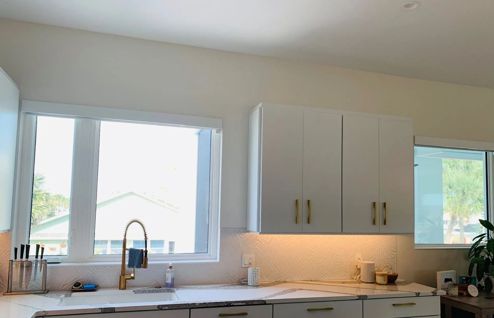 White kitchen with cabinets, sink, and window. Gold faucet and hardware. Sunlight streams in.