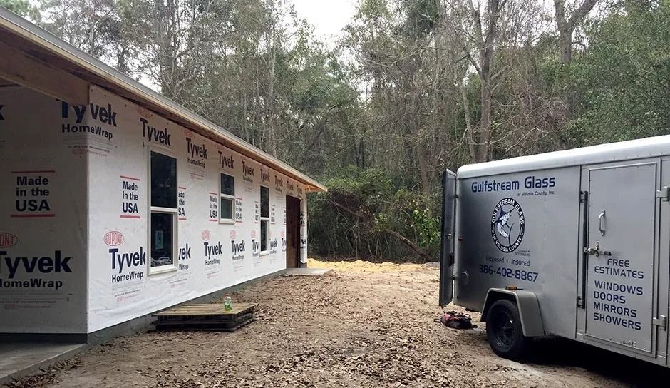 A building wrapped in Tyvek with windows, a trailer, and a wooded backdrop.