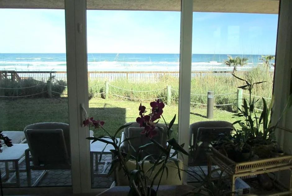 View of ocean from inside, through a window. Green grass, beach, blue sky and water. Plants inside.
