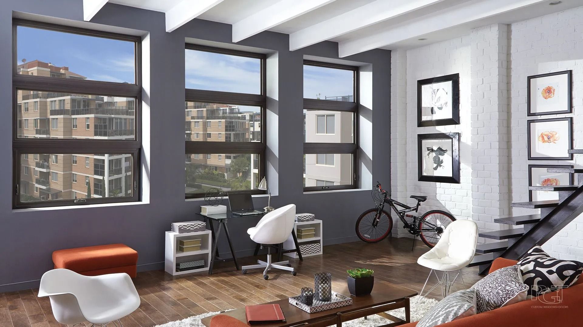Modern living room with tall windows, desk, bike, and orange accents against gray walls.