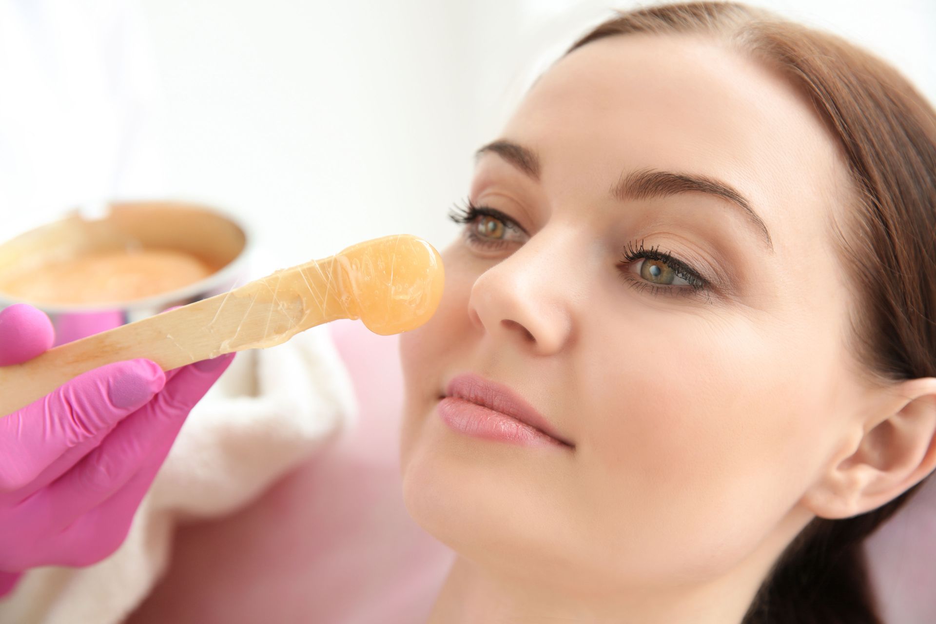 Woman getting facial hair waxed, pink gloved hand holding wax applicator.