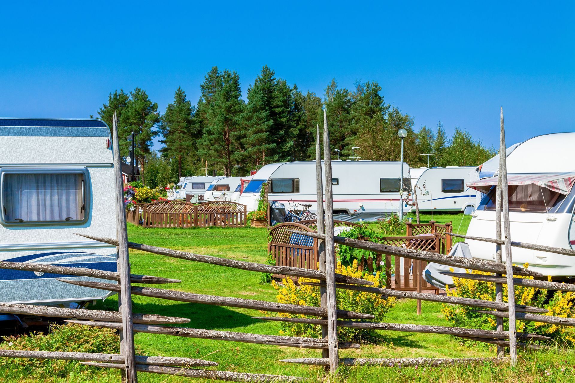 Campground with RVs parked on green grass, wooden fences, and trees under a clear blue sky.