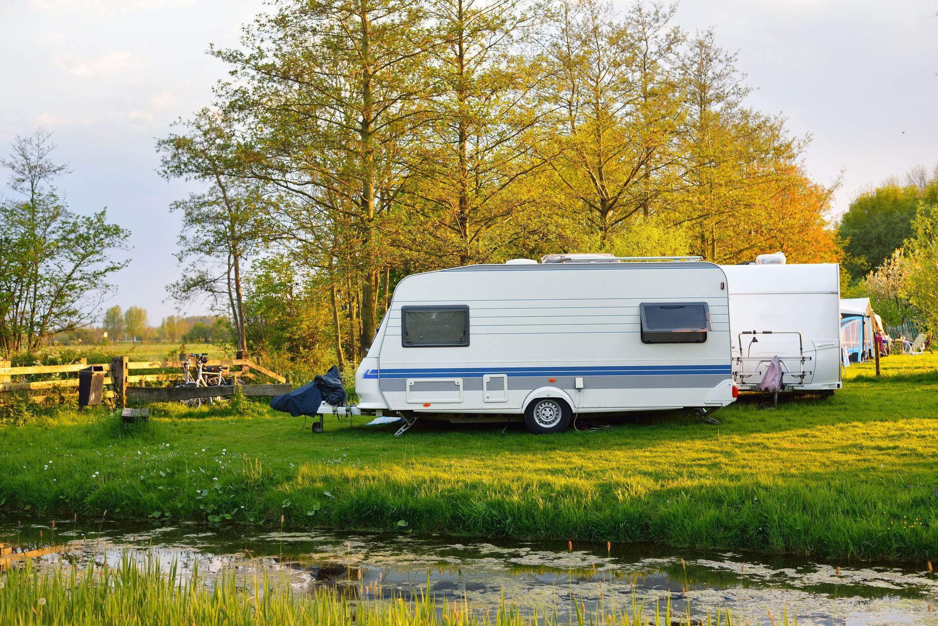 Camping trailers parked on a grassy field near trees and a small body of water.
